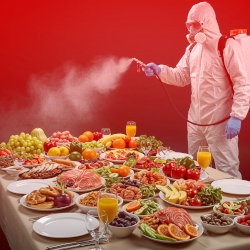 A man in a biohazard suit spraying a fine mist over a table full of food.